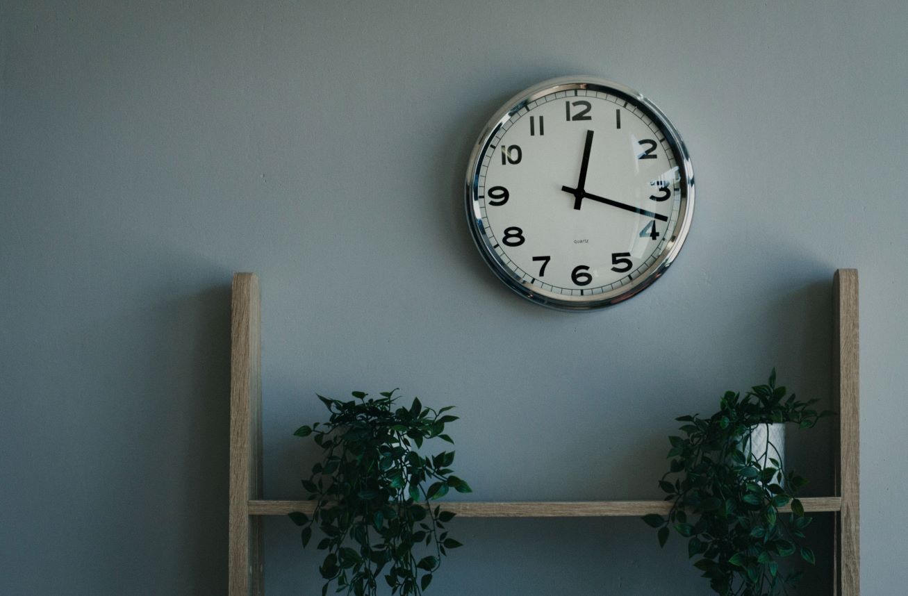 Relógio numa parede com estante embaixo e plantas representando o post sobre o que significa asap em inglês